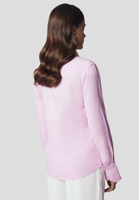 Jasnoróżowa koszula z lnu z kołnierzykiem, długimi rękawami i mankietami na guziki. Posiada zaokrąglony dół i luźny krój, zaprezentowana od tyłu.