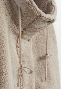 Lys beige fleecejakke med høj krave, der har justerbare snore og en tekstureret, blød overflade.