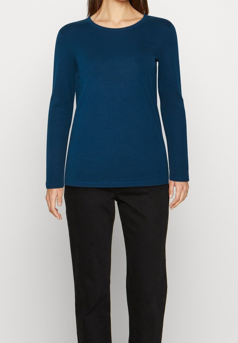 Top de manga larga en tono teal, confeccionado en tejido suave, con cuello redondo y corte ajustado, combinado con pantalones negros. Textura suave y diseño minimalista.