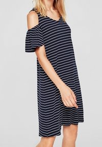 Kvinna som bär en marinblå och vit horisontellt randig klänning med kalla axlar och korta, fladdrande ärmar mot en enfärgad bakgrund.