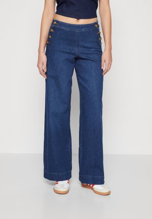 Vrouw draagt een high-waisted donkerblauwe wijde broekspijpspijkerbroek met decoratieve knopen op de heupen en witte sneakers met rode strepen.