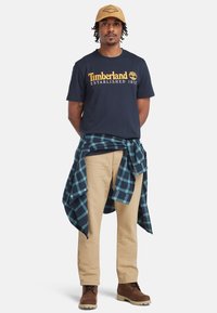 Námořnické tričko s žlutým logem, béžové kalhoty, hnědé boty. Modro-zelená kostkovaná košile uvázaná kolem pasu. Model stojí s pažemi podél těla.