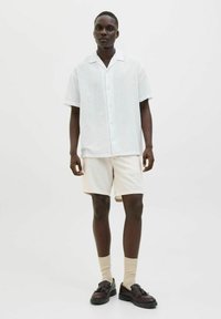 Camisa blanca de manga corta y ligera con cuello, combinada con pantalones cortos beige. Zapatos marrones y calcetines beige completan el atuendo. Diseño simple y casual.