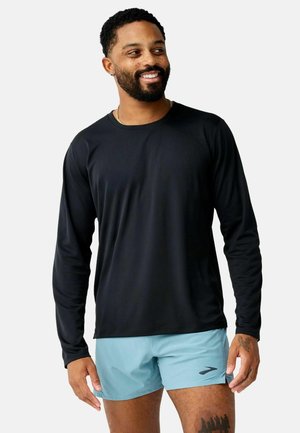 Uomo con barba che indossa una maglietta nera a maniche lunghe e pantaloncini azzurri, sorridente e che guarda di lato su uno sfondo uniforme.