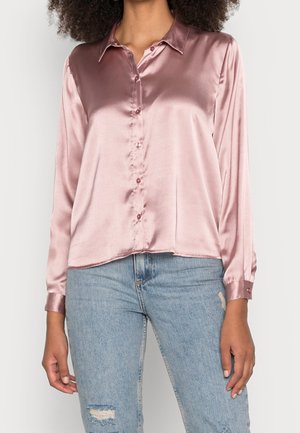 Kobieta w jasno-różowej, satynowej koszuli z guzikami, w połączeniu z jasnoniebieskimi, podartymi dżinsami, stojąca na jednolitym tle.