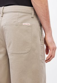 Bézs színű pamut nadrág hátsó zsebbel, apró piros Calvin Klein logó címkével. Sima textúra, szabott kialakítás és klasszikus fazon.