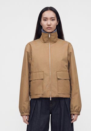 Frau mit langen schwarzen Haaren trägt eine beige Reißverschlussjacke mit hohem Kragen und zwei vorderen Klappentaschen über dunklen, weit geschnittenen Hosen.