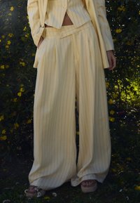 Světlé žluté pruhované sako s dvojřadým zapínáním a širokými kalhotami. Jemná textura s výraznými záhyby a odpovídajícími knoflíky.