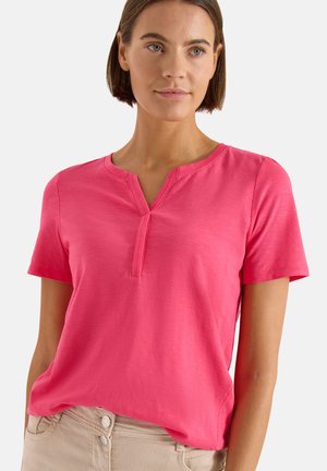 Kvinde iført en knaldrød kortærmet skjorte med en delt halsudskæring, let stoppet ned i beige bukser med to synlige knapper.