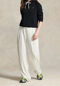 Černá mikina s kapucí a bílými šňůrkami, spojená s volnými bílými plisovanými kalhotami a multikolorovými teniskami s černými a žlutými akcenty.