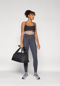 Czarny stanik sportowy z logo "NEBBIA", szare legginsy o wysokim stanie i szare sneakersy, trzymając czarną torbę sportową z krótkimi uchwytami.