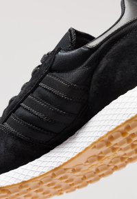 Musta tennari, jossa mokkanahkainen ja teksturoidusta kankaasta valmistettu päällinen, joustavat yksityiskohdat sekä valkoinen välipohja, jossa teksturoitu kumipohja.