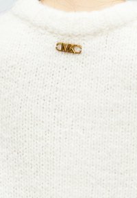 Pull en tricot blanc avec un col rond et une épingle logo MK dorée à l'arrière. La texture est douce avec un motif de tricot serré.