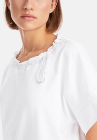 Weißes kurzärmliges Oberteil mit Rüschenausschnitt und Kordelzugdetails. Glatte Textur, lockere Passform und ohne Muster, die den Fokus auf Minimalismus legt.