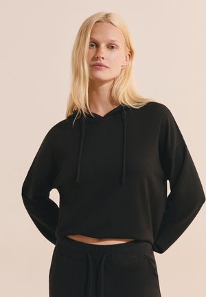 Femme blonde portant un sweat à capuche court noir ample et un pantalon noir avec une ceinture, debout les mains derrière le dos.