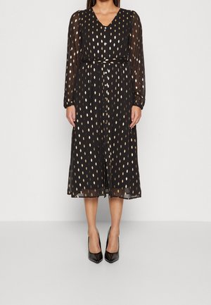 Žena v černých midi šatech se zlatými kapkovitými vzory, průsvitnými dlouhými rukávy, páskem v pase a černými lodičkami s špičatou špičkou stojící na bílém pozadí.