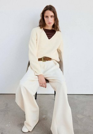 Femme assise sur une chaise, portant un pull crème par-dessus un top rayé marron, un pantalon large crème et une ceinture marron avec une boucle dorée.