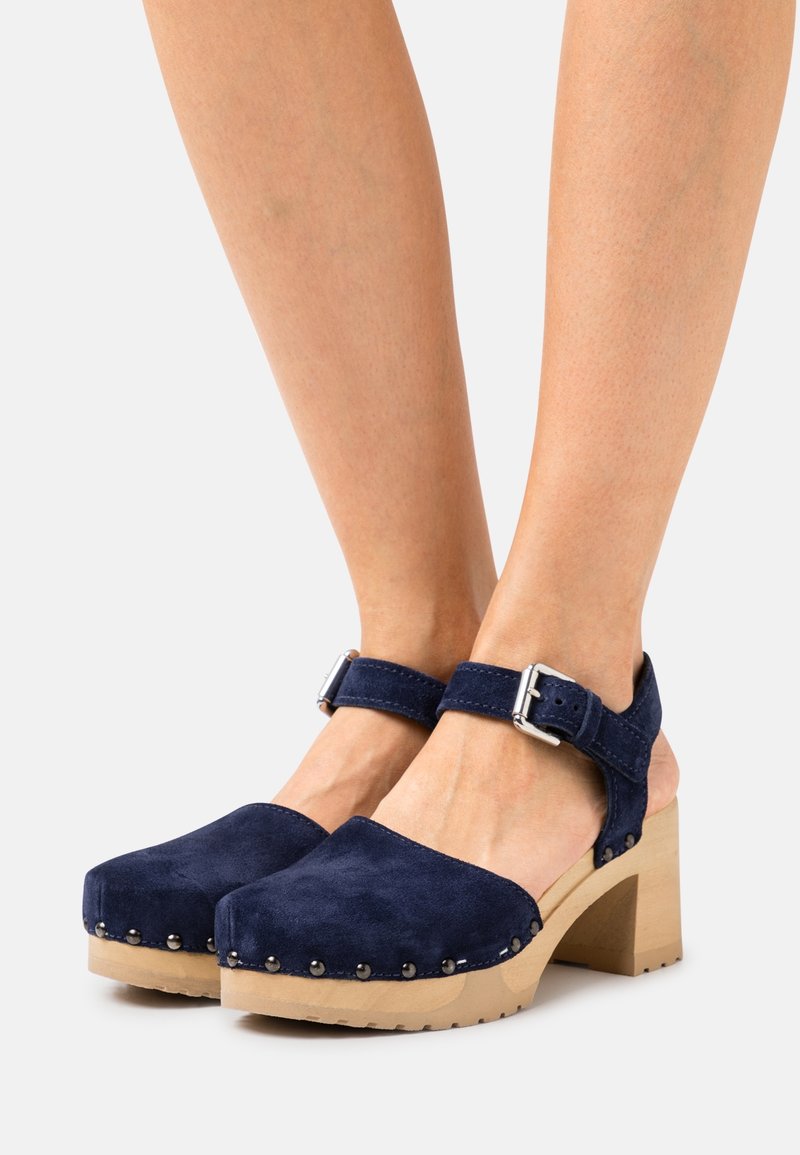 Sabots en daim bleu marine avec talons en bois épais, attachés par des brides à la cheville avec des boucles argentées, portés pieds nus.