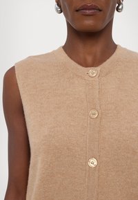 Beige strikket ermeløs topp med rund hals, tre knapper foran og en myk tekstur. Enkel design uten mønstre.