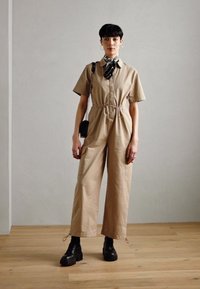 Beige jumpsuit with short sleeves, drawstring waist, side pockets, paired with chunky black boots and a patterned scarf. Wood floor backdrop.