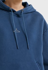 Felpa con cappuccio blu realizzata in tessuto morbido, con cordoncino con punte scure e un sottile logo a forma di appendiabiti ricamato sul petto.