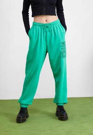 Osoba nosząca turkusowe spodnie dresowe Nike z elastycznymi mankietami oraz czarne buty na platformie, stojąca na zielonej podłodze na białym tle.