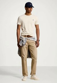 Man wearing beige t-shirt, khaki pants, white sneakers, navy cap, plaid shirt tied around waist, standing on plain light background.