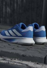 Chaussures de sport bleues avec des bandes blanches et des semelles blanches, placées côte à côte sur une surface pavée sur un fond sombre.