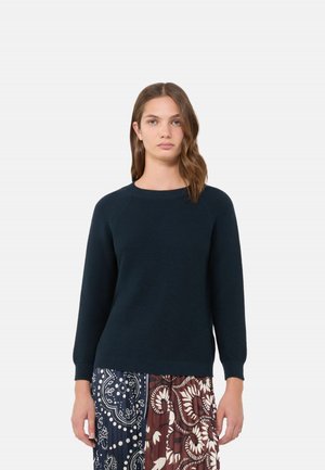 Donna con capelli castani fino alle spalle che indossa un maglione scuro a maniche lunghe e una gonna con motivi blu navy e marrone.