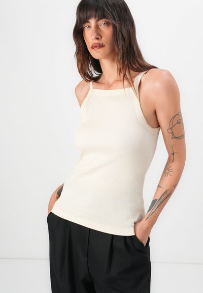 Lichtbeige geribde tanktop met een vierkante halslijn en dunne bandjes. De stof is glad met een aansluitende silhouette. Draagt met zwarte broek.