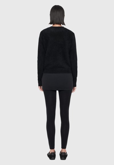 Vue de dos d’une personne portant un pull noir, une jupe courte, un legging et des baskets sur un fond clair uni.