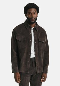 Bruine suède shirtjack met een kraag, twee borstzakken, drukknoopsluitingen en een gladde textuur, gedragen over een wit t-shirt.