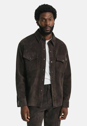 Brown suede shirt jacket with a collar, two chest pockets, snap buttons, and a smooth texture, worn over a white t-shirt.