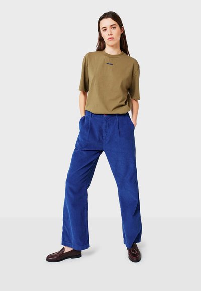 Donna in piedi con le mani in tasca, che indossa una maglietta verde oliva, pantaloni in velluto a coste blu larghi e mocassini marroni scuri su uno sfondo grigio.