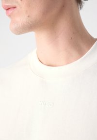 Cream cotton t-shirt with a ribbed crew neckline and embossed "HUGO" logo on the front. Smooth texture and casual fit.