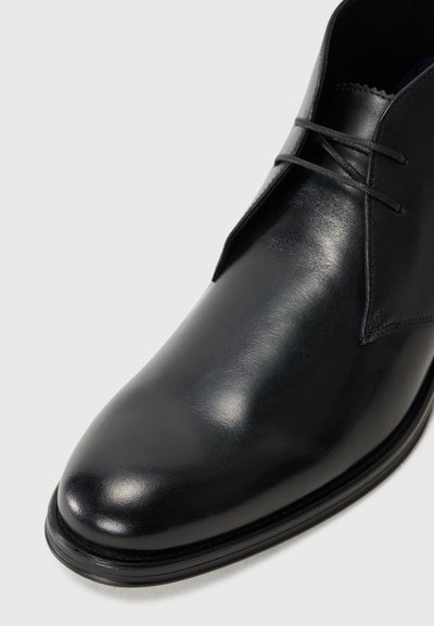 Botte chukka en cuir noir au fini lisse, à bout arrondi, au design minimaliste, dotée de lacets et de subtils accents de couture.