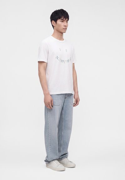 Νέος άνδρας στέκεται φορώντας λευκό graphic t-shirt, ανοιχτό μπλε τζιν και λευκά αθλητικά παπούτσια σε απλό φωτεινό φόντο.