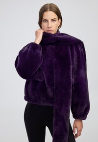 Donna che indossa un giubbotto di pelliccia morbida di un viola scuro e una sciarpa abbinata, con pantaloni neri, in piedi contro uno sfondo chiaro e uniforme.