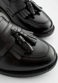 Chaussures habillées en cuir noir avec des accents en gland, texture lisse, bout arrondi et des coutures contrastantes le long de la semelle.