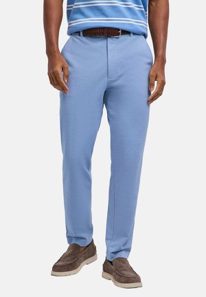 Uomo che indossa pantaloni azzurri, cintura marrone, camicia a righe blu e mocassini in camoscio marrone, in piedi su uno sfondo bianco.