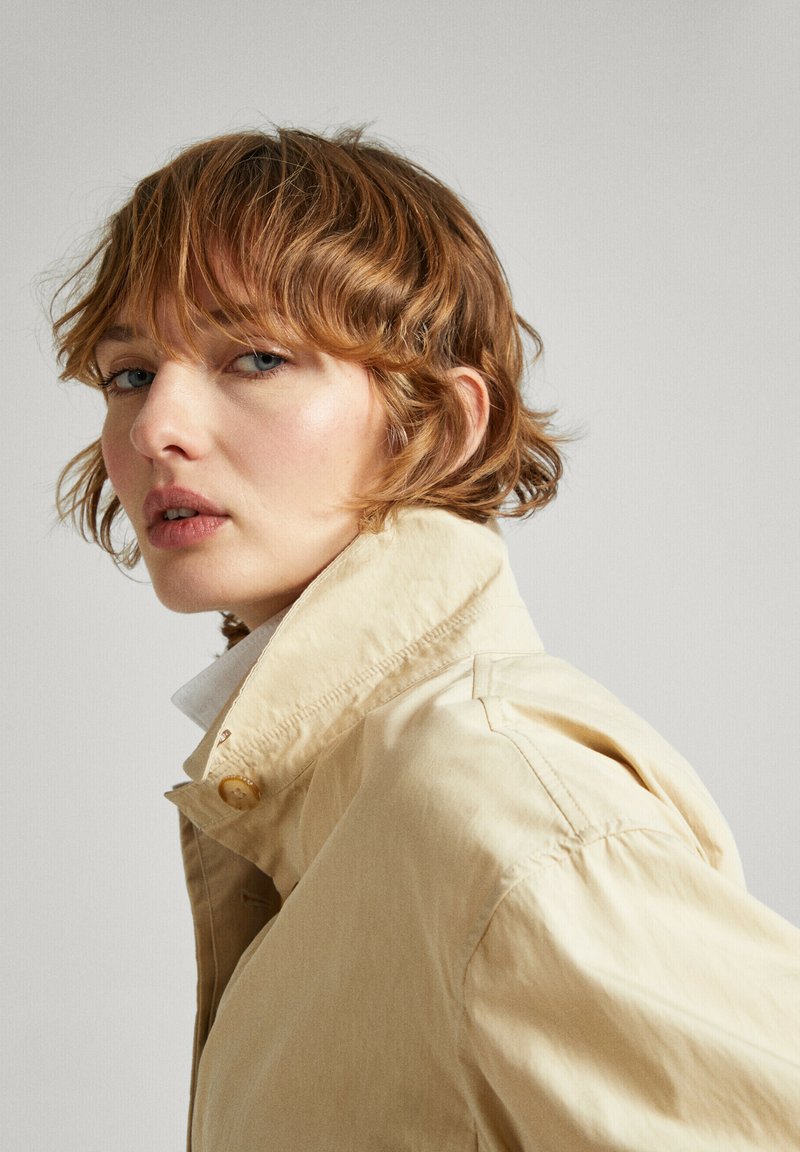 Veste beige légère à col avec des détails de boutons et un tissu texturé, offrant une coupe décontractée et des accents de couture visibles.