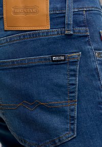 Vaqueros de denim en un color azul oscuro con un acabado texturizado. Las características incluyen costuras naranjas, una etiqueta de cuero etiquetada "BIG STAR" y una pequeña etiqueta en el bolsillo trasero.