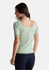 Světle zelený top s krátkým rukávem, s vykrojeným výstřihem na zádech, přiléhavý střih a hladká textura. Jednobarevný, bez vzorů nebo ozdob, v kombinaci s tmavými džíny.
