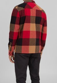 Punainen ja musta ruutuhame flanellikuvioinen paita vaaleanbeigeillä yksityiskohdilla, jossa on nappikaulus ja säädettävät napit hihansuissa, esitetty takaa.