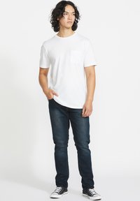 Camiseta blanca de algodón con un bolsillo en el pecho, combinada con jeans de mezclilla oscuros. El modelo está de pie de manera casual con las manos en los bolsillos, usando zapatillas negras.