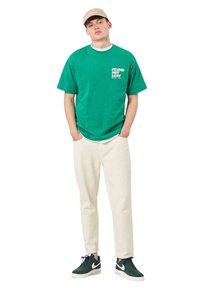 Joven de pie con las manos en los bolsillos, vistiendo una camiseta verde con el texto "FOUND NOT LOST", pantalones beige, zapatillas oscuras y una gorra beige.