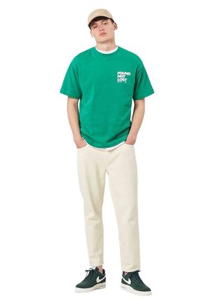 Giovane uomo in piedi con le mani nelle tasche, indossa una maglietta verde con la scritta "FOUND NOT LOST", pantaloni beige, scarpe da ginnastica scure e un cappellino beige.