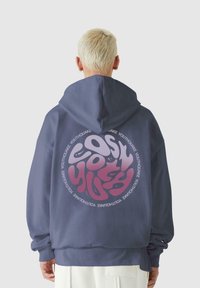 Tumši zils kapučinš ar lielu apaļu grafiku aizmugurē. Grafika satur stilizētu tekstu rozā un baltā krāsā. Mīksta tekstūra, brīva piegriezuma.