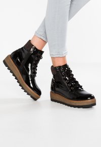 Bottines en cuir verni noir avec une semelle plateforme texturée beige, lacets à l'avant et fermeture éclair sur le côté. Semelle extérieure en caoutchouc avec motif en zigzag.