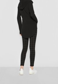 Černý dlouhý svetr s kapucí a zakřiveným lemem, který je doplněn černými legínami s bílou mřížkovou strukturou a bílými teniskami.